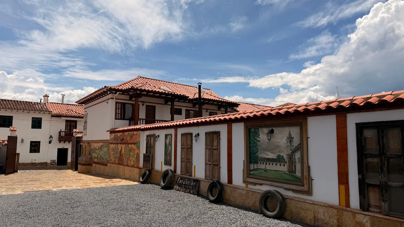 Parqueadero de la posada con piso de gravilla, casona blanca de techos de teja y muros con murales y pinturas